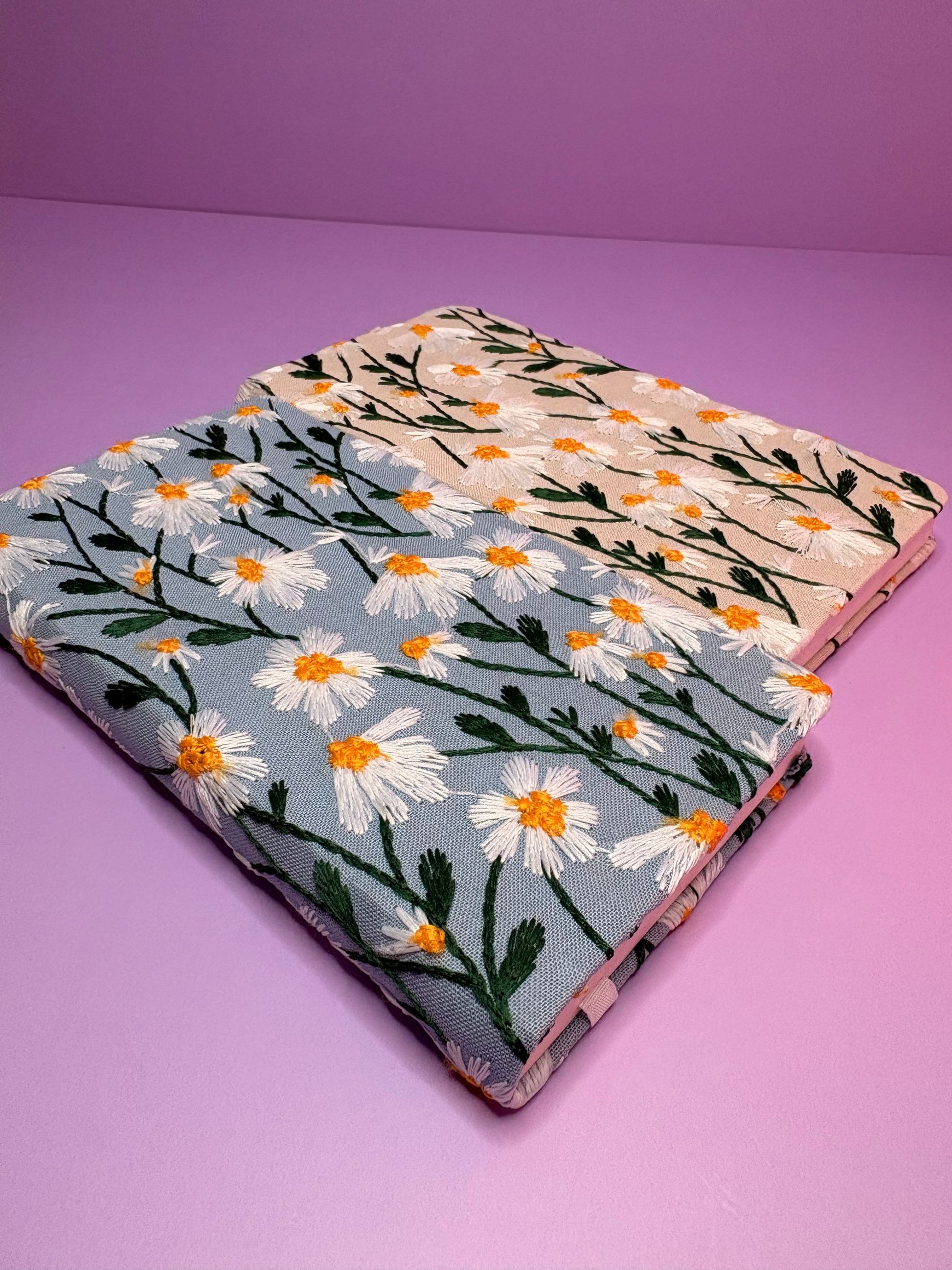 Two A6 embroidered daisy notebooks with hardback covers in blue and cream on a purple background