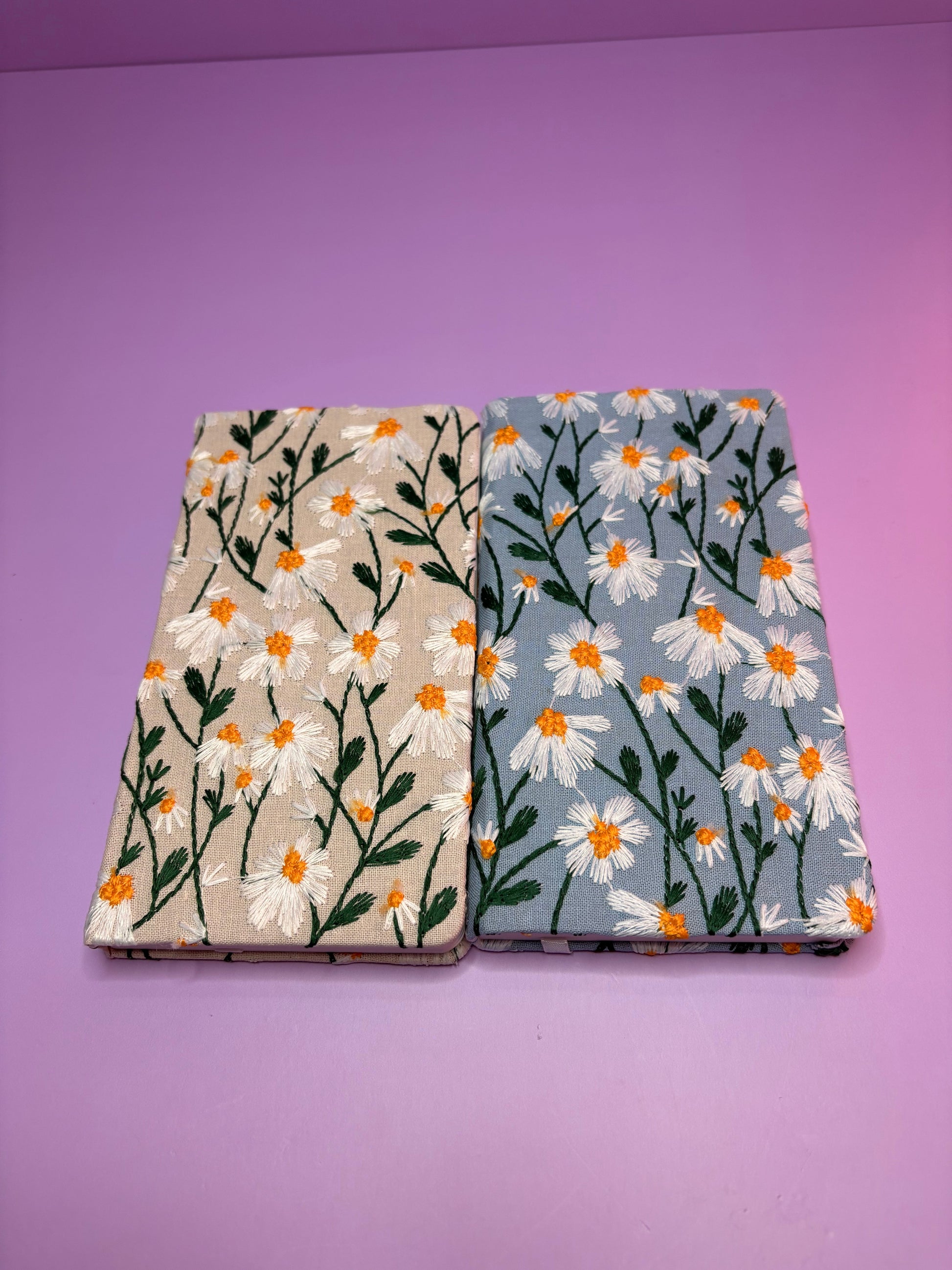 Two A6 embroidered daisy notebooks with cream and blue covers on a purple background