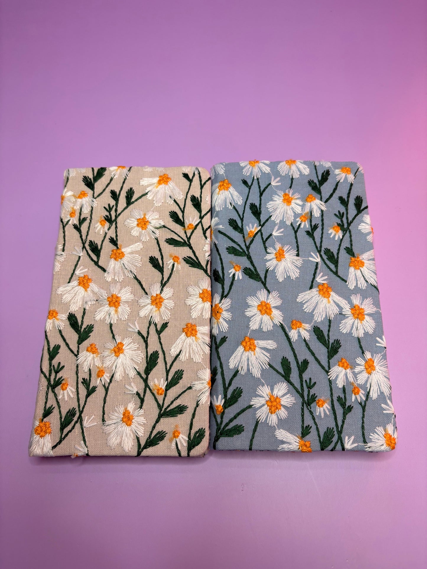 Two A6 embroidered daisy hardback notebooks in blue and cream covers on a purple background
