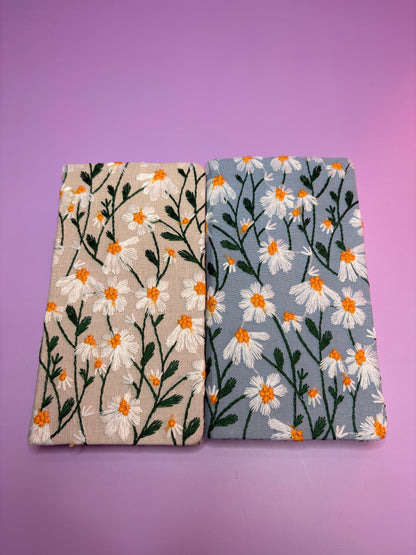Two A6 embroidered daisy hardback notebooks in blue and cream covers on a purple background
