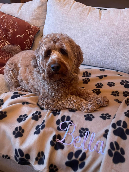 Personalised dog blanket with paw prints and embroidered name, featuring a fluffy dog resting on it.