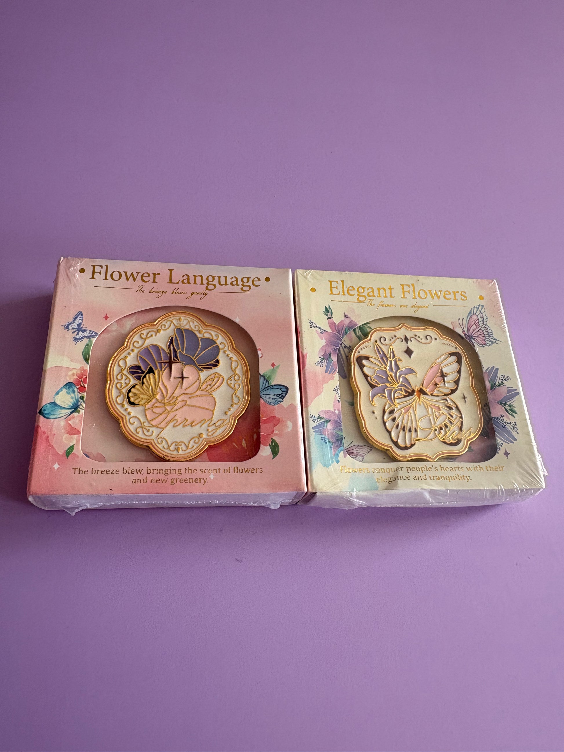 Two decorative flower-shaped items in their retail packaging, placed side by side on a solid purple background. The left box is labeled 'Flower Language' and is pink with a floral design, while the right box is labeled 'Elegant Flowers' and is cream with butterfly and floral artwork.