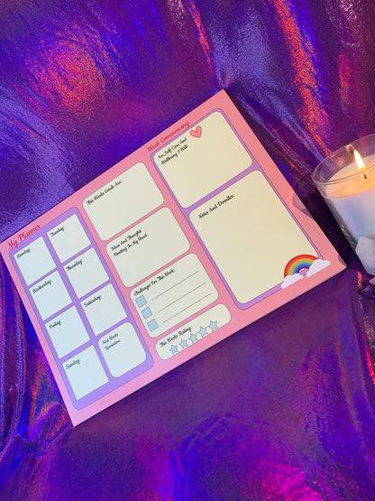 The Pretty in Pink A4 Desk Planner with illustrated pages on a shiny purple surface, accompanied by a lit candle.